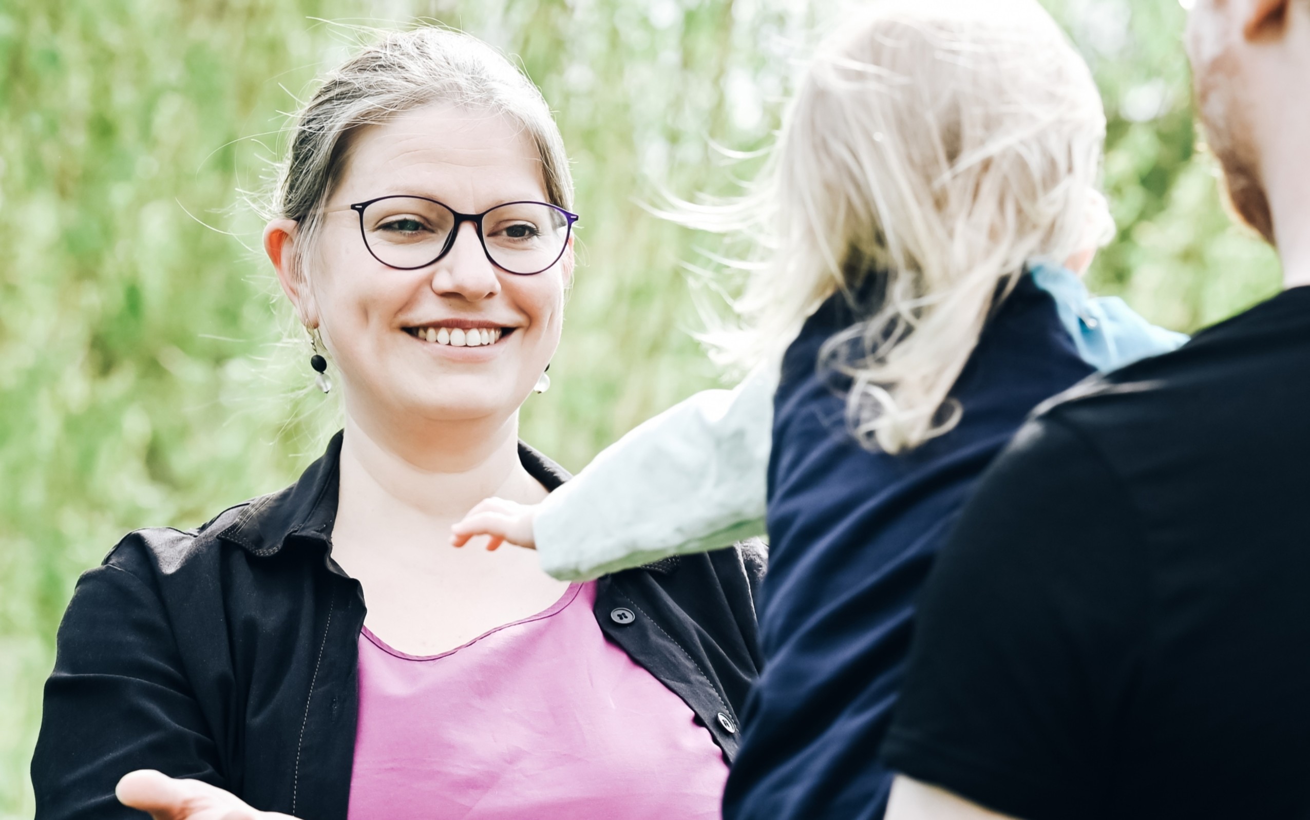 Ronja steht lächelnd mit ausgebreiteten Armen und nimmt ein Kind mit blonden Haaren entgegen.